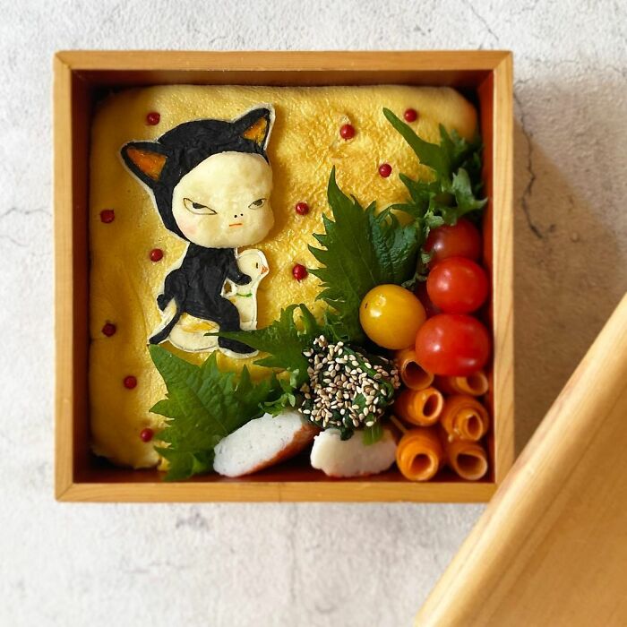 Bento artist crafting detailed lunchbox design featuring character art with fresh vegetables and sesame seeds in wooden box.