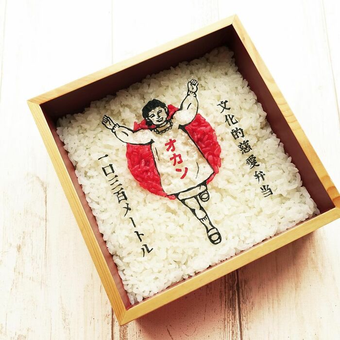 Bento artist creates detailed lunchbox featuring character art and Japanese text on a bed of white rice with red background.