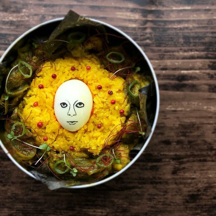 Bento artist lunchbox featuring a creative face-drawn egg surrounded by yellow rice and greens on wooden surface.