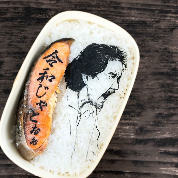 Bento artist creates detailed lunchbox featuring a character sketch and Japanese text on rice beside cooked salmon.