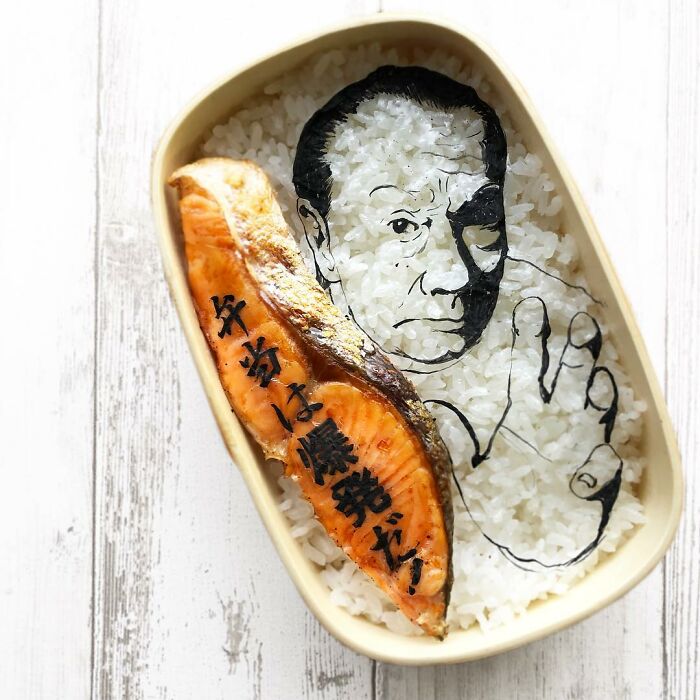 Bento artist creates creative lunchbox with salmon and detailed rice art of a man's face and hand gesture.