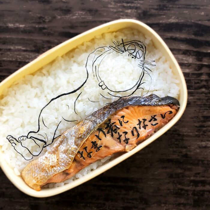 Bento artist’s creative lunchbox with salmon and a manga character drawn over rice for a personalized meal.