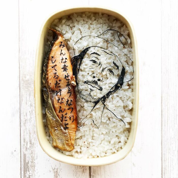 Bento artist creates detailed lunchbox art featuring a character sketch and Japanese text on rice with a piece of grilled fish.