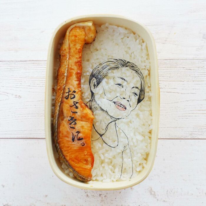 Bento art lunchbox featuring a detailed face illustration on rice and a grilled fish piece with Japanese characters.