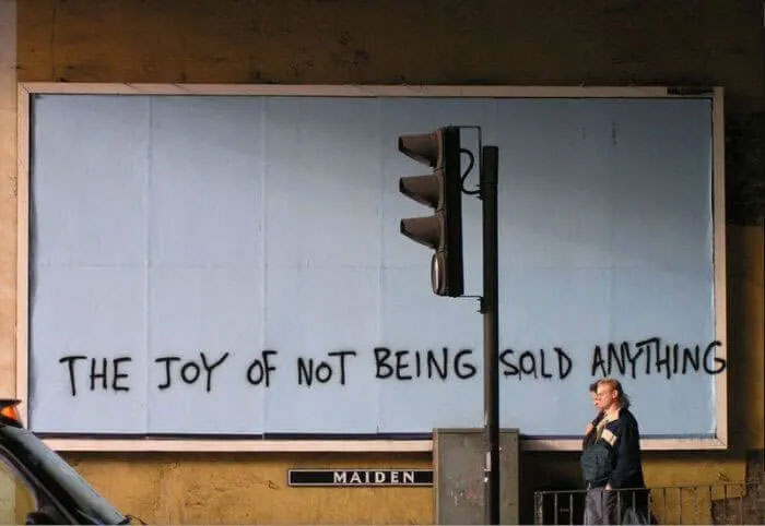 Graffiti on a billboard reads the joy of not being sold anything, illustrating anti-consumption ideas in a street setting.