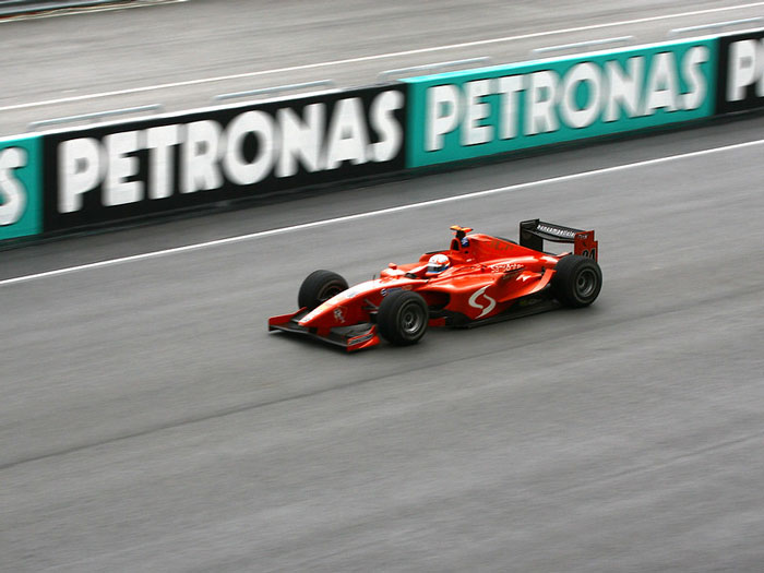 Red Formula 1 race car speeding on a track with Petronas sponsorship banners in the background.