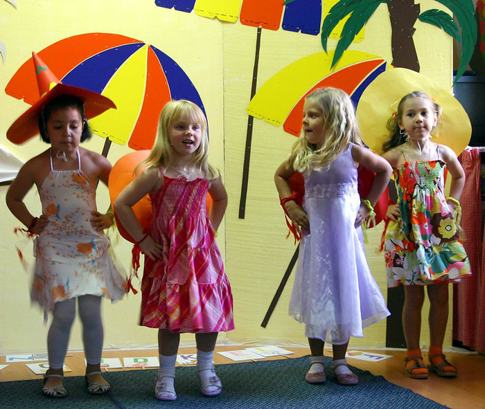 Four young girls in colorful dresses and festive hats performing on stage with decorations in an online group setting.