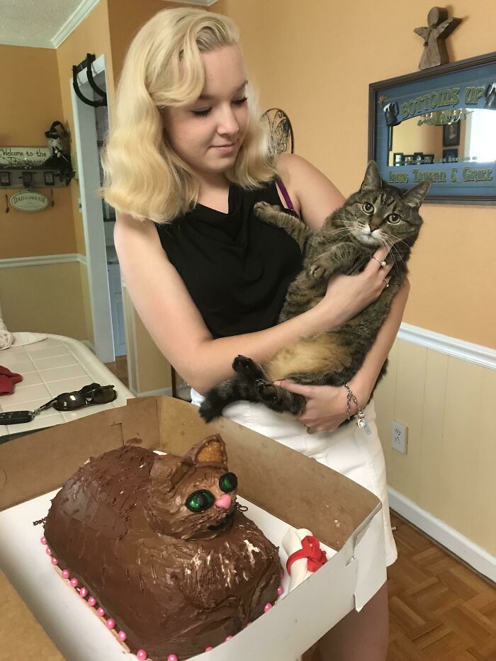 Woman holding cat next to a humorous cake fail resembling a cat with green eyes and pink decorations.