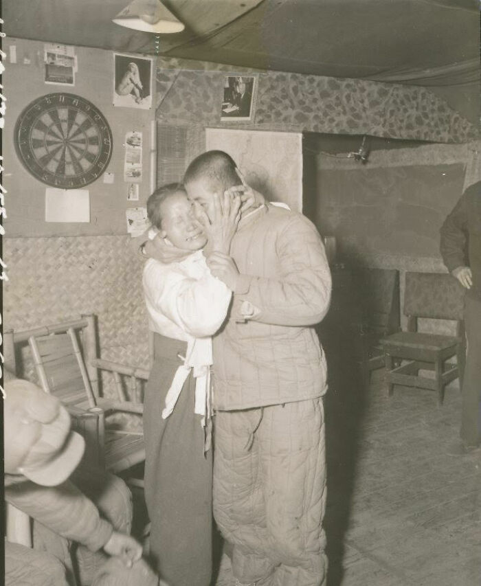 A soldier and a woman embrace in a room adorned with a dartboard and photos, capturing a historical moment.