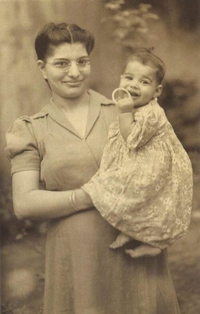 A historical photo of a smiling woman holding a baby, both dressed in vintage clothing, capturing a moment of joy.