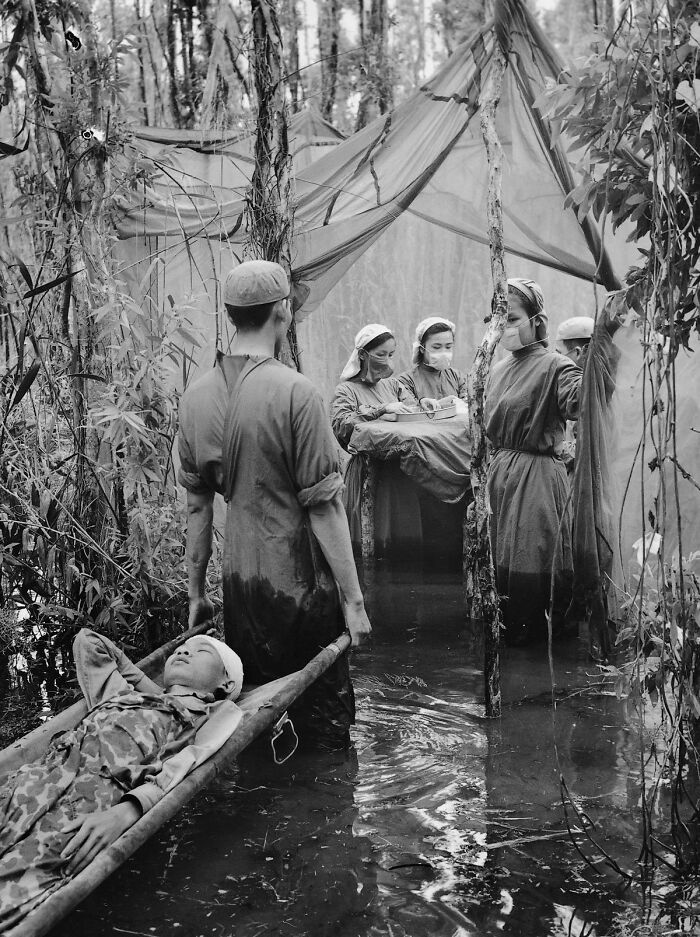 Historical photo of medics in a flooded forest treating a patient on a stretcher under a makeshift shelter.
