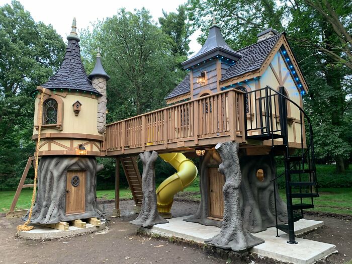 Intricately crafted woodworking treehouse with towers, slide, and detailed railings in a wooded backyard setting.