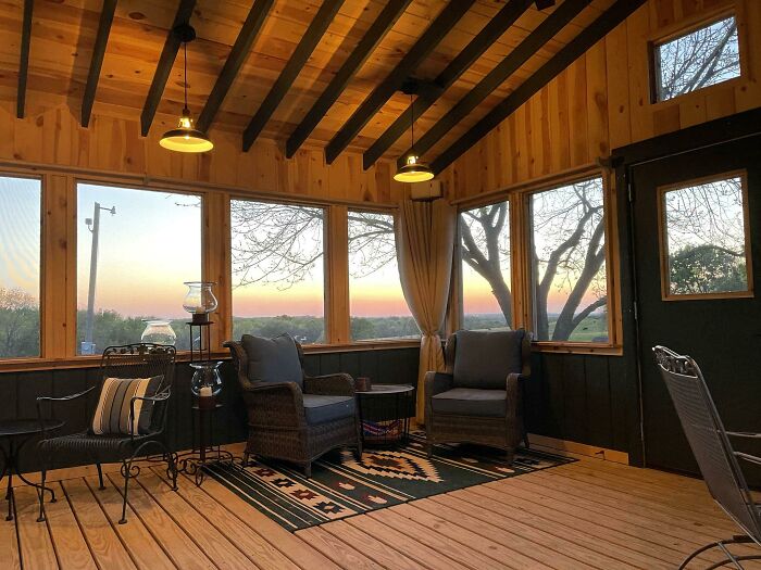 Cozy woodworking porch with wooden floors, beams, chairs, and a scenic sunset visible through large windows.