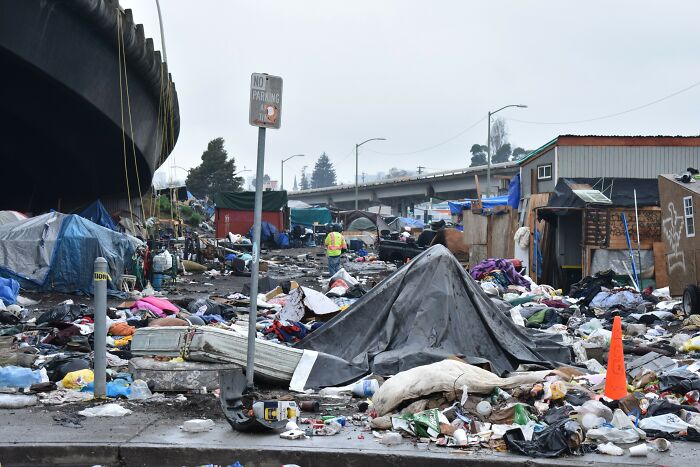 Urban hell scene with homeless encampment, scattered trash, and makeshift shelters under highway overpass in dystopian city setting.