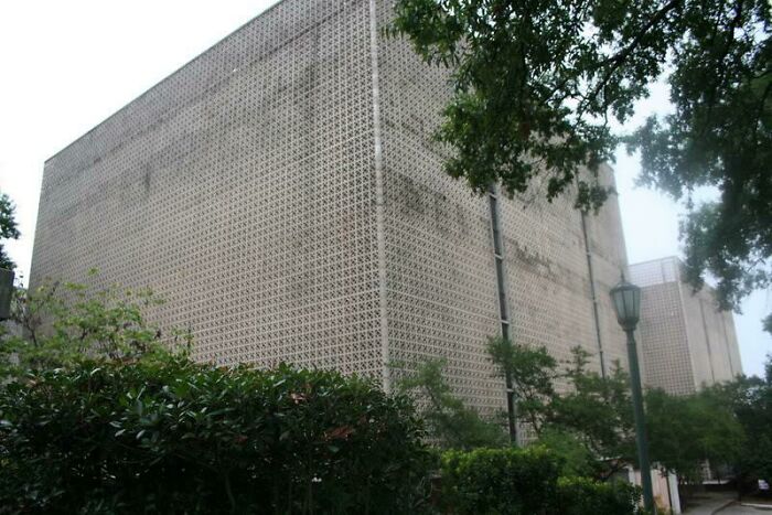 Large concrete building with repetitive patterns surrounded by overgrown greenery in an urban hell dystopian scene.