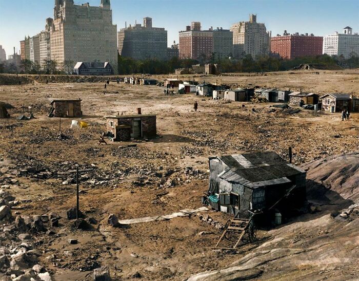 Dystopian urban hell scene with rundown shacks and empty barren land near tall apartment buildings.