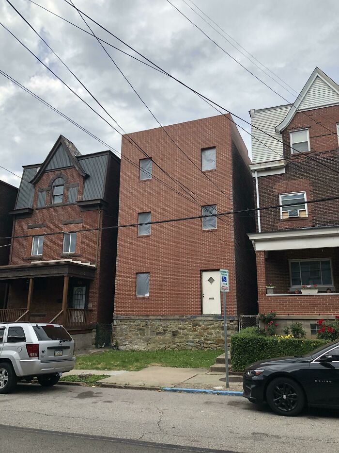 Brick building with boarded-up windows between two older houses, depicting urban hell and dystopian real-life scene.