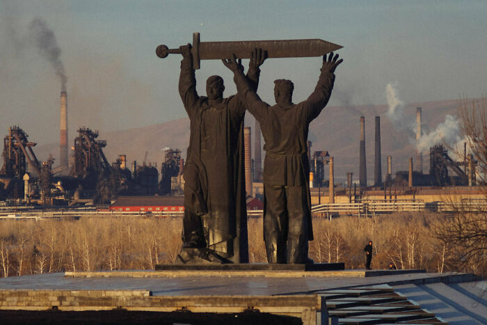 Statues holding a large sword with industrial factories emitting smoke in the background, depicting urban hell dystopia.