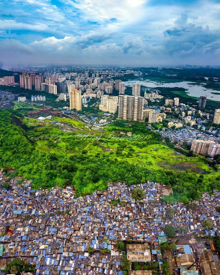 Aerial view of urban hell showing sprawling slums contrasted with high-rise buildings and green spaces under a cloudy sky.