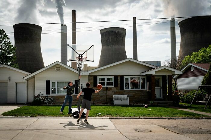 Two boys playing basketball outside a house with large industrial cooling towers in the background in urban hell setting.