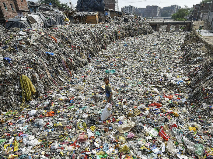 Child standing amid vast urban hell of garbage-filled drainage channel in a densely populated area, showing dystopian reality.