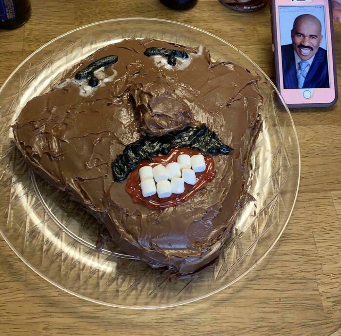 Funny cake fail with a chocolate-frosted face and marshmallow teeth, next to a phone displaying a reference image.