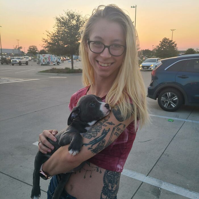 Young woman smiling and holding a black and white puppy in a parking lot at sunset, rescue pet pics concept.