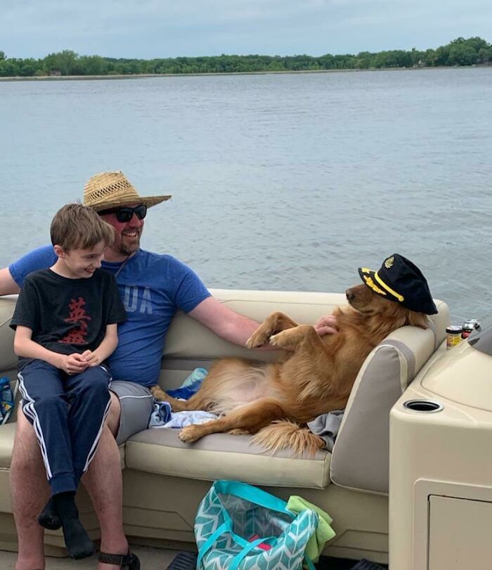 Dog acting bizarre wearing a captain's hat, lounging on a boat couch beside smiling people by the water.