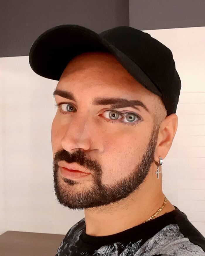 Man with a beard and black cap featuring optical illusion body art that creates convincing eye effects on his face.