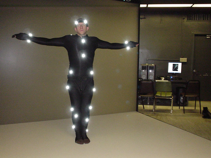 Person in motion capture suit with reflective markers standing with arms outstretched in a studio setting for online group.