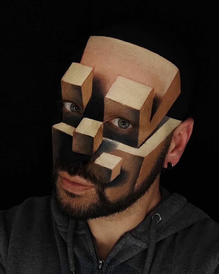 Italian artist with body paint creating a convincing optical illusion of 3D geometric shapes on his face against a black background.