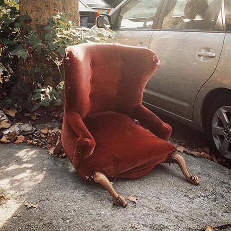 Old red armchair with missing legs placed oddly on a sidewalk next to parked cars, illustrating weird Instagram account content.