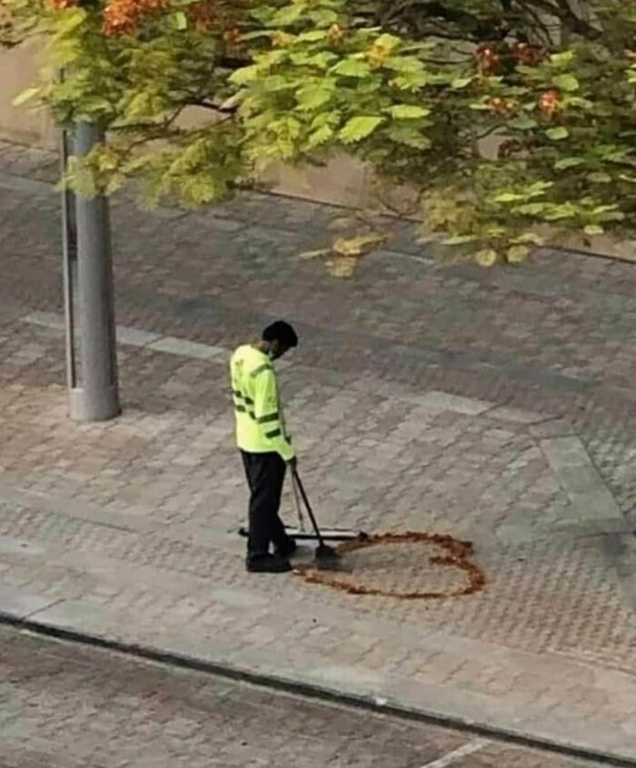 Street worker in a neon jacket sweeping leaves shaped like a heart on a sidewalk, weird Instagram account content.