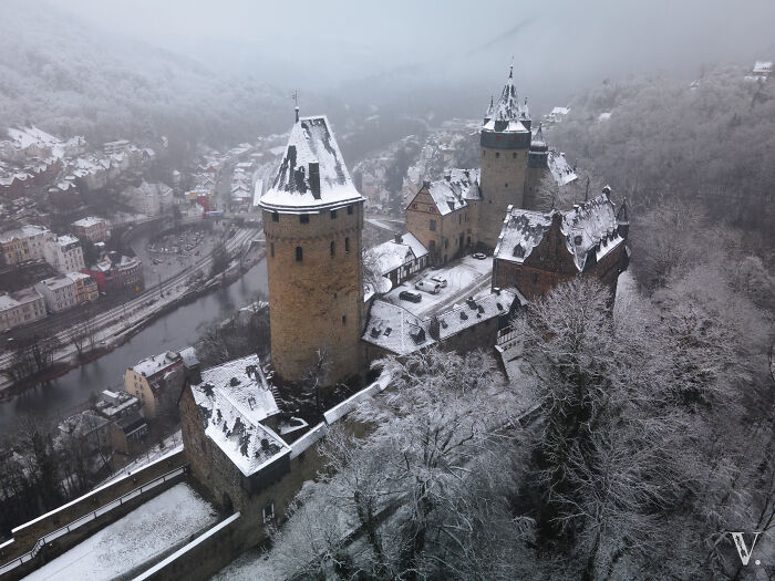Aerial view of a beautiful medieval castle covered in snow surrounded by fog and winter trees along a river valley.