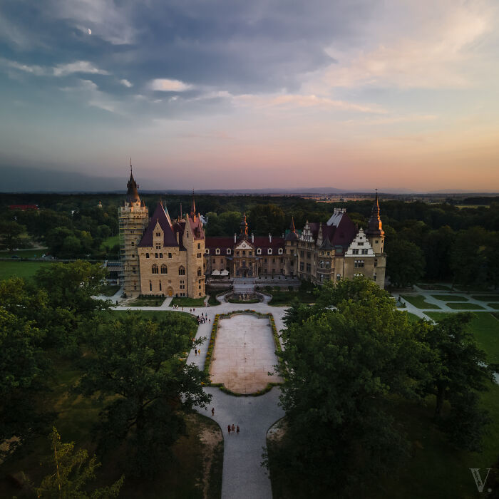 Aerial view of a beautiful castle surrounded by trees and gardens under a colorful sunset sky.