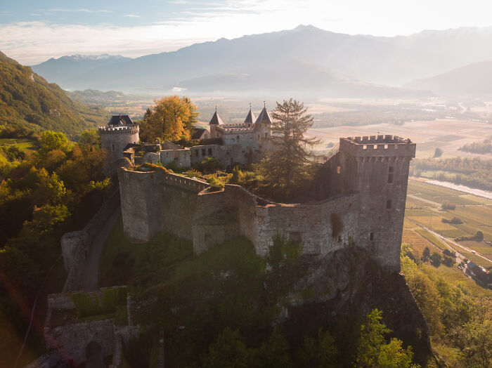 Ancient stone castle surrounded by trees on a hill with mountains and fields in the background at sunset.