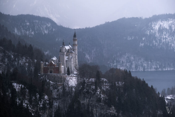 Scenic view of a beautiful castle surrounded by snowy mountains and dense forest in a misty winter landscape.