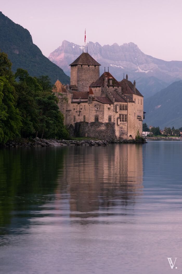 Historic castle by calm lake with mountain backdrop at sunset, showcasing one of the most beautiful castles around the world.