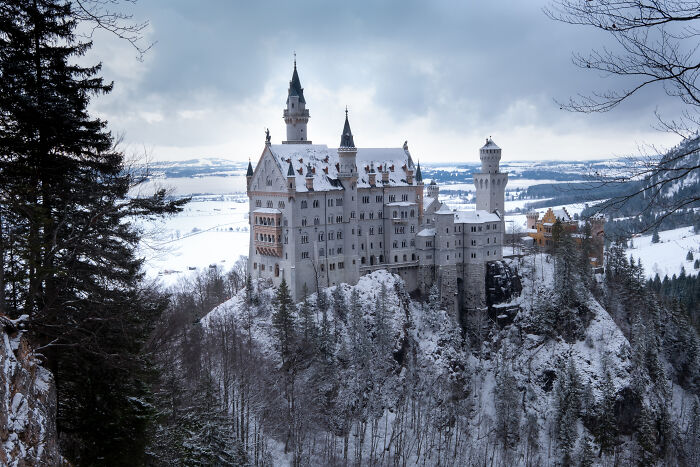 Snow-covered beautiful castle perched on a rocky cliff surrounded by winter forest and mountains in a scenic landscape.