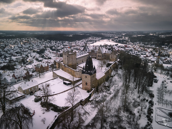 Aerial view of a beautiful snow-covered castle surrounded by a town and forest under a dramatic cloudy sky.
