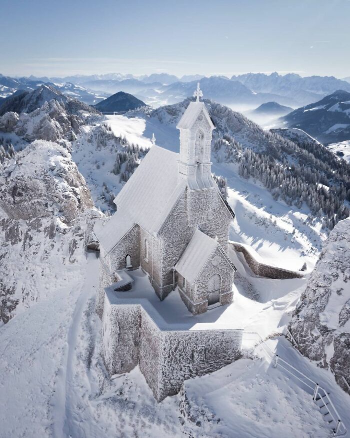 Snow-covered castle on a mountain surrounded by snowy trees and distant mountain ranges under clear sky.