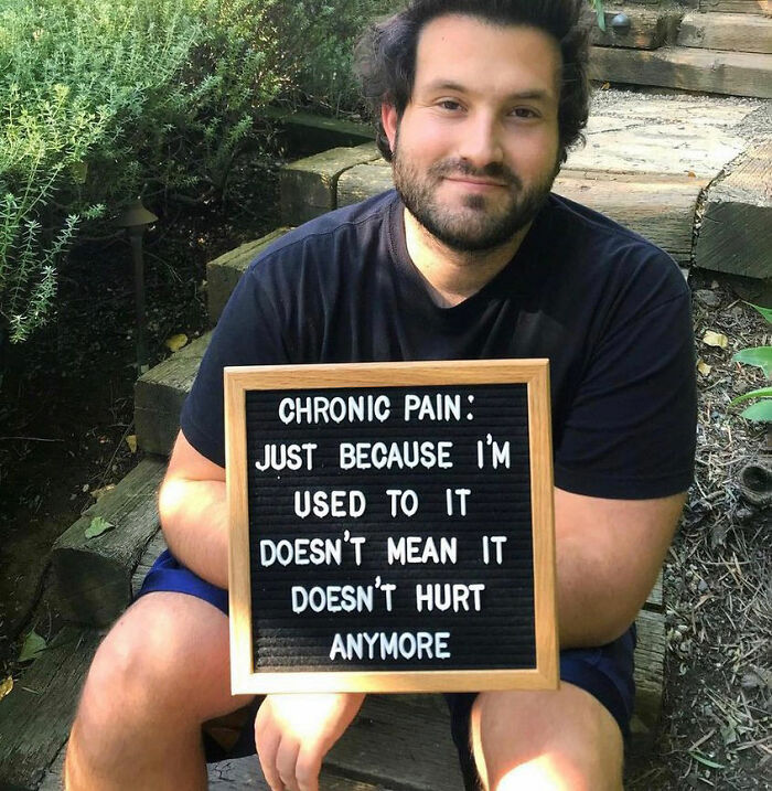 Man sitting outdoors holding a sign about chronic pain, raising awareness of challenges faced by people with chronic illnesses.