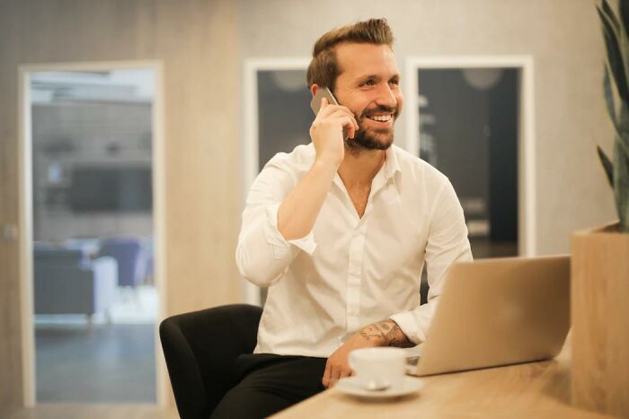 Smiling man in white shirt using phone at modern office desk with laptop, showing subtle things about wealthy people.