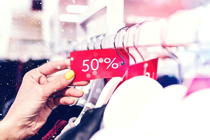 Hand with yellow nails holding a 50 percent off tag on clothing rack showing subtle signs of wealthy people shopping habits.