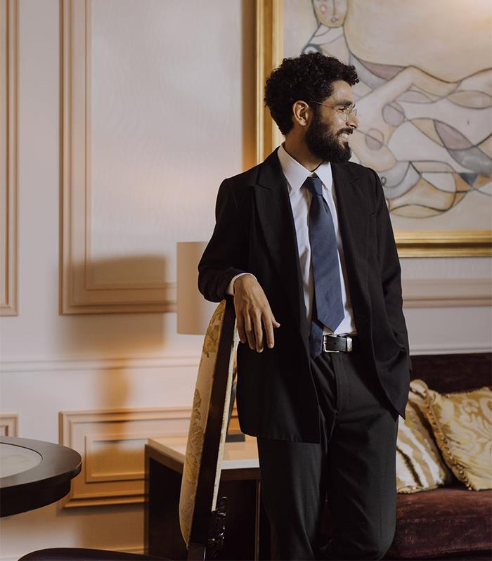 Man in a suit and tie leaning on a chair in an elegant room, representing subtle signs of wealthy people.