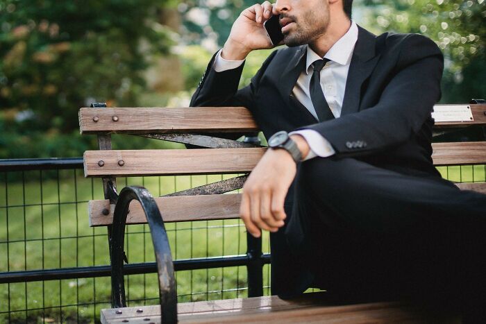 Man in a black suit on a bench speaking on phone, showcasing subtle signs of wealthy people and rich lifestyle.