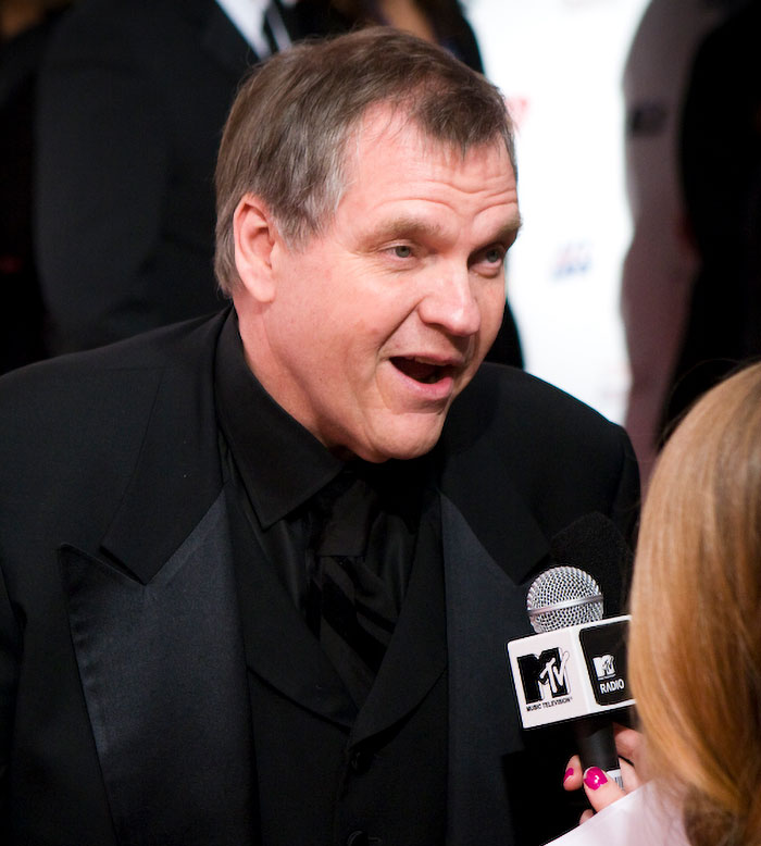 Middle-aged man in black suit being interviewed with MTV microphone, related to taxi and Uber drivers sharing crazy stories.