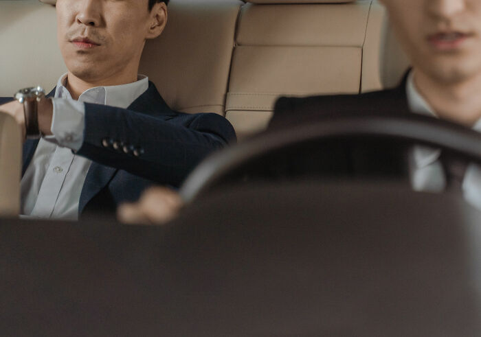 Two men in a car, one driving and one sitting in the back, representing taxi and Uber drivers on the job.