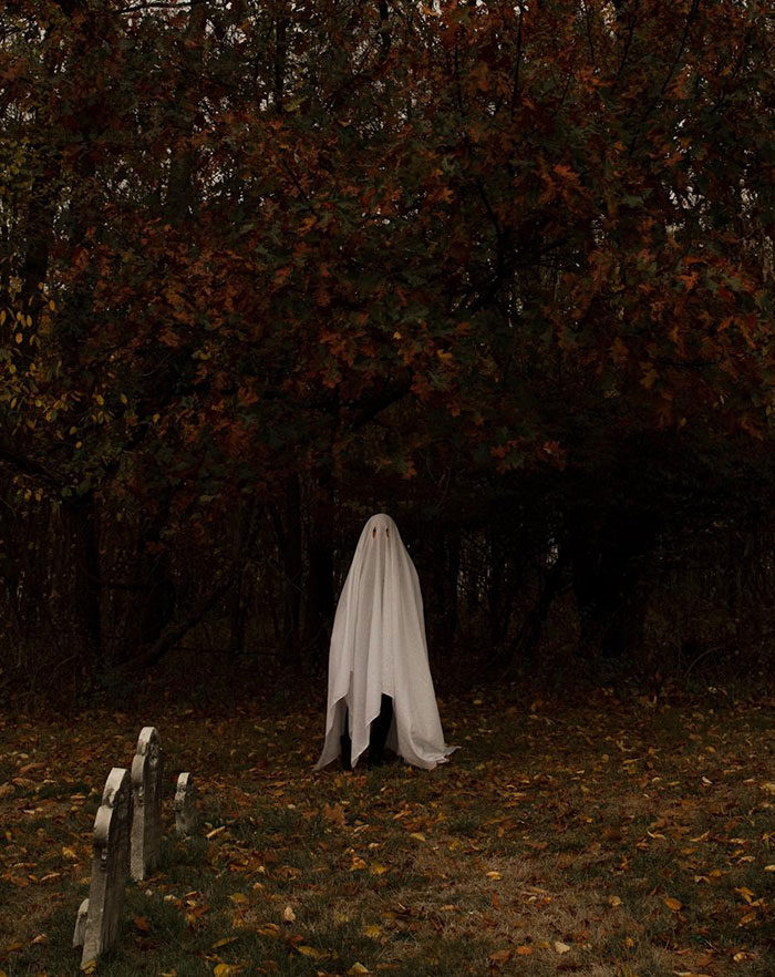 Person in a simple ghost costume made with a white sheet standing in a dark outdoor area with trees and gravestones nearby.