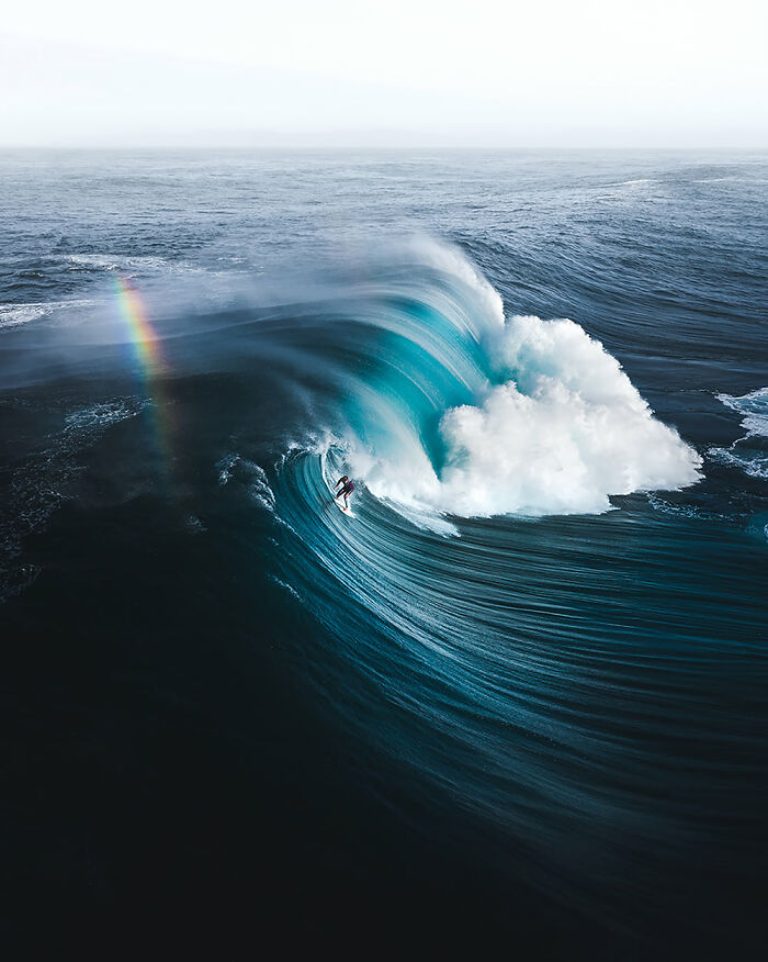 Surfer riding a large ocean wave with a visible rainbow in mist captured for ocean photography awards finalists.